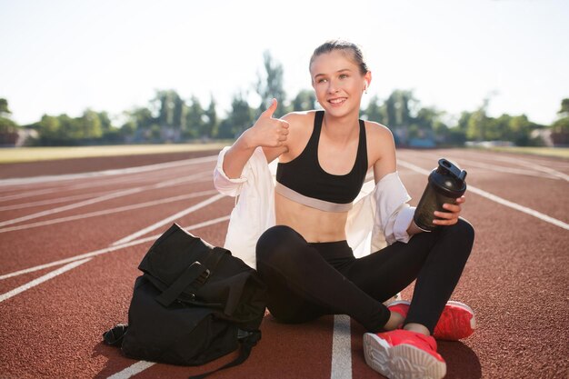 Ragazza gioiosa in auricolari wireless che tiene in mano una bottiglia sportiva che guarda felicemente da parte mentre mostra il pollice in su sulla pista da corsa dello stadio