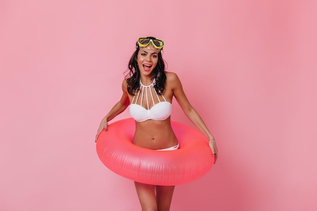 Ragazza gioiosa in bikini in piedi su sfondo rosa Donna bruna sorridente in posa con cerchio di nuoto