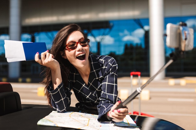 Ragazza gioiosa in occhiali da sole con mappa appoggiata su un'auto cabriolet mentre scatta felicemente foto sul cellulare con passaporto in mano