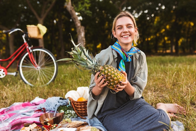 Ragazza gioiosa seduta su una coperta da picnic sull'erba che tiene felicemente l'ananas in mano trascorrendo del tempo nel parco
