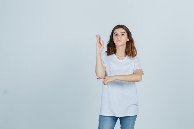 Ragazza giovane alzando la mano in maglietta bianca e guardando fiducioso, vista frontale.