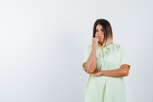 Ragazza giovane appoggiando il mento sul pugno, tenendo la mano sotto il gomito in maglietta e guardando pensieroso, vista frontale.