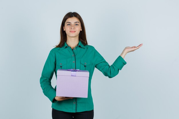 Ragazza giovane azienda confezione regalo, allargando il palmo in camicetta verde, pantaloni neri e guardando felice. vista frontale.