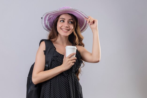 Ragazza giovane bella viaggiatore in cappello estivo con zaino tenendo la tazza di caffè sorridente allegramente felice e positivo in piedi su sfondo bianco