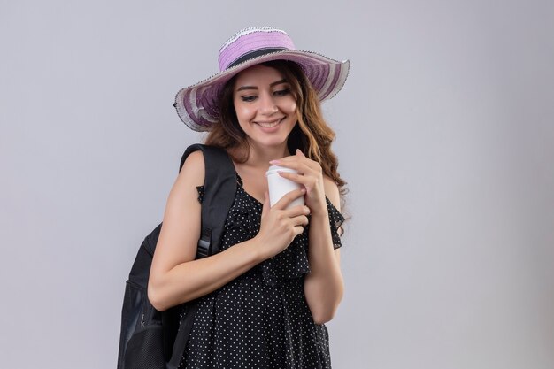 Ragazza giovane bella viaggiatore in cappello estivo con zaino tenendo la tazza di caffè sorridente allegramente felice e positivo in piedi su sfondo bianco