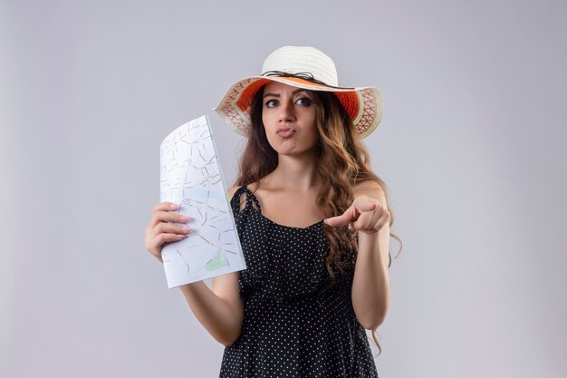 Ragazza giovane bella viaggiatore in vestito a pois in cappello estivo che punta con il dito alla fotocamera con espressione scettica sul viso in piedi su sfondo bianco