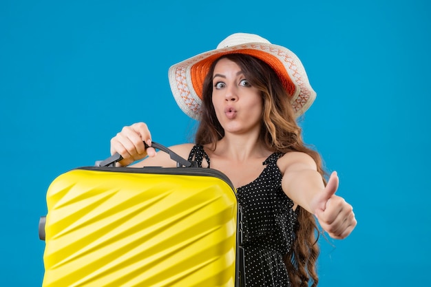 Ragazza giovane bella viaggiatore in vestito a pois in cappello estivo che tiene la valigia guardando uscito e felice che mostra i pollici in su gioendo del suo successo e vittoria in piedi su sfondo blu