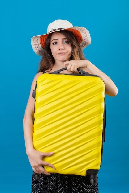 Ragazza giovane bella viaggiatore sconvolto in vestito a pois in cappello estivo che tiene la valigia che guarda l'obbiettivo con espressione triste sul viso in piedi su sfondo blu