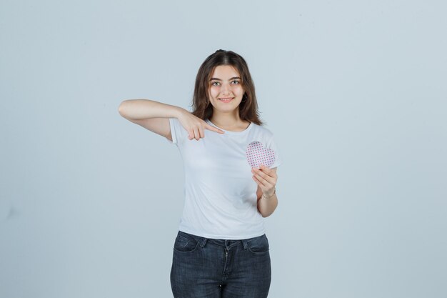 Ragazza giovane che indica il contenitore di regalo mentre lo si tiene in t-shirt, jeans e guardando allegro, vista frontale.