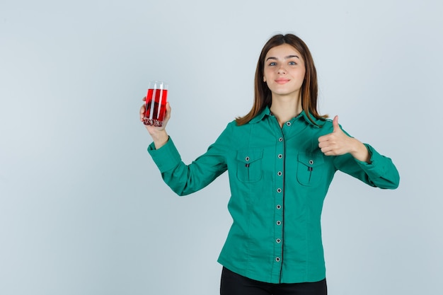 Ragazza giovane con un bicchiere di liquido rosso, mostrando il pollice in su in camicetta verde, pantaloni neri e guardando felice. vista frontale.