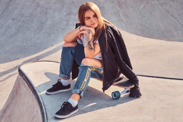 Ragazza giovane e bella skater vestita alla moda seduta sul longboard allo skatepark.
