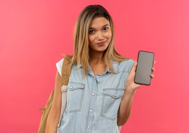 Ragazza giovane e carina studentessa lieta che indossa la borsa posteriore che mostra il telefono cellulare nella parte anteriore isolata sulla parete rosa