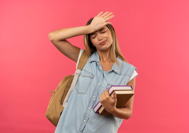 Ragazza giovane e carina studentessa stanca che indossa la borsa posteriore che tiene i libri che mettono la mano sulla fronte con gli occhi chiusi isolati su fondo rosa con lo spazio della copia