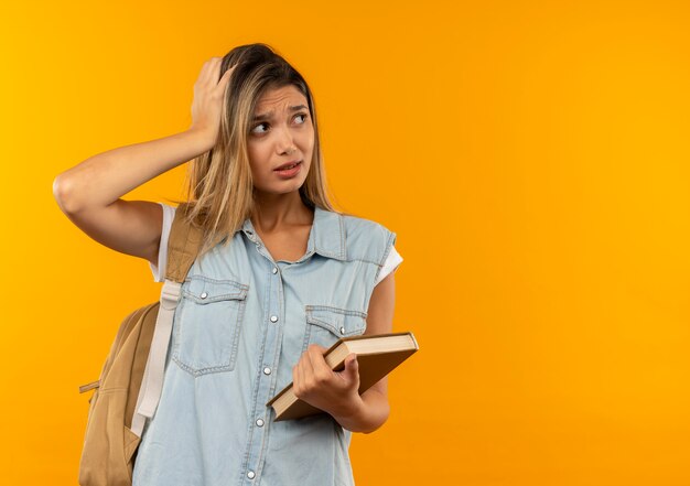 Ragazza giovane e graziosa dell'allievo confusa che porta il libro posteriore della tenuta della borsa che mette la mano sulla testa che esamina il lato isolato sull'arancia con lo spazio della copia