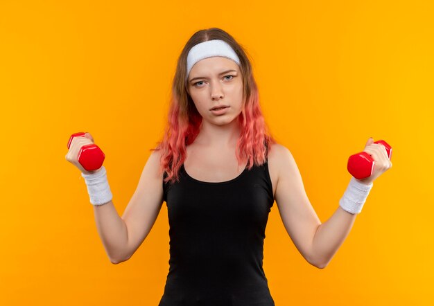 Ragazza giovane fitness in abiti sportivi utilizzando manubri facendo esercizi cercando fiducioso in piedi sopra la parete arancione