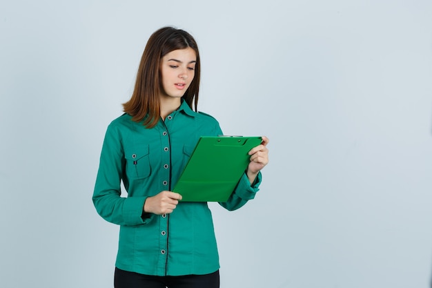 Ragazza giovane guardando appunti in camicetta verde, pantaloni neri e guardando concentrato, vista frontale.