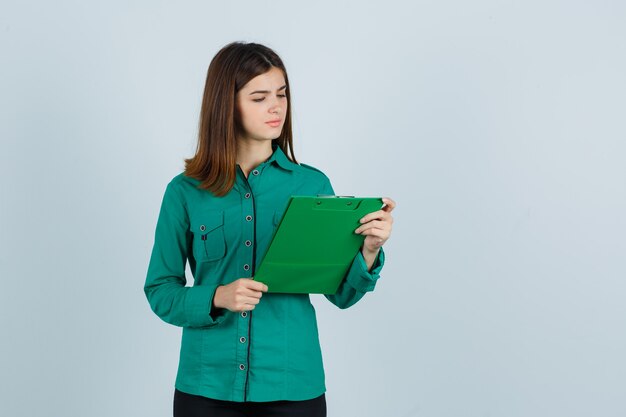 Ragazza giovane guardando appunti in camicetta verde, pantaloni neri e guardando confuso, vista frontale.