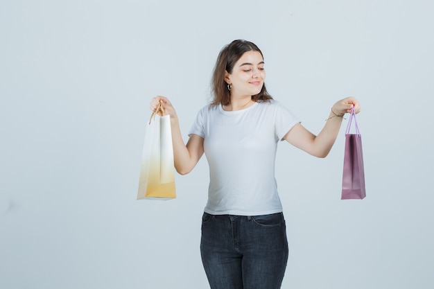 Ragazza giovane mantenendo i sacchetti regalo in t-shirt, jeans e guardando soddisfatto, vista frontale.