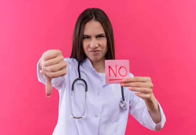 Ragazza giovane medico spiacevole che indossa abito medico stetoscopio tenendo nessuna nota il pollice verso il basso sul muro rosa isolato