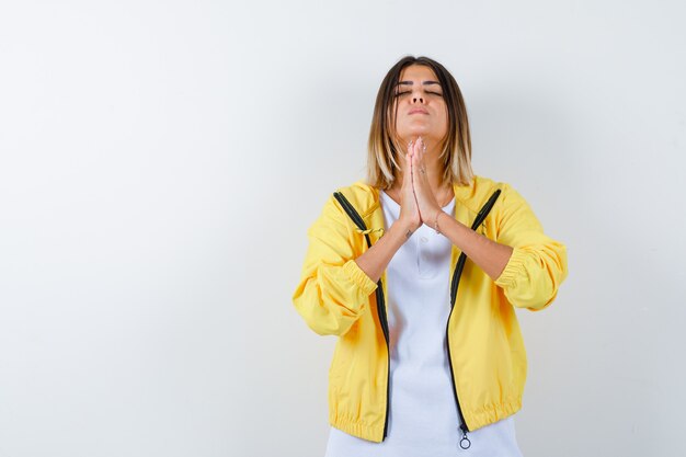 Ragazza giovane stringendo le mani in posizione di preghiera in maglietta bianca, giacca gialla e guardando concentrato, vista frontale.