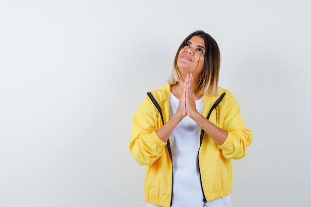 Ragazza giovane stringendo le mani in posizione di preghiera in t-shirt bianca, giacca gialla e guardando allegro, vista frontale.