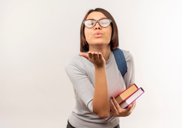 Ragazza giovane studente con gli occhiali e borsa posteriore che tengono i libri che inviano il bacio del colpo alla macchina fotografica isolata su fondo bianco con lo spazio della copia