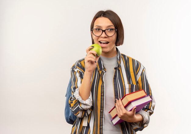 Ragazza giovane studente con gli occhiali e borsa posteriore che tengono i libri e si prepara a mordere la mela isolata su priorità bassa bianca con lo spazio della copia