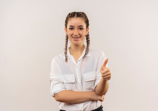 Ragazza giovane studente con le trecce in camicia bianca guardando in avanti sorridente che mostra i pollici in su in piedi sul muro bianco