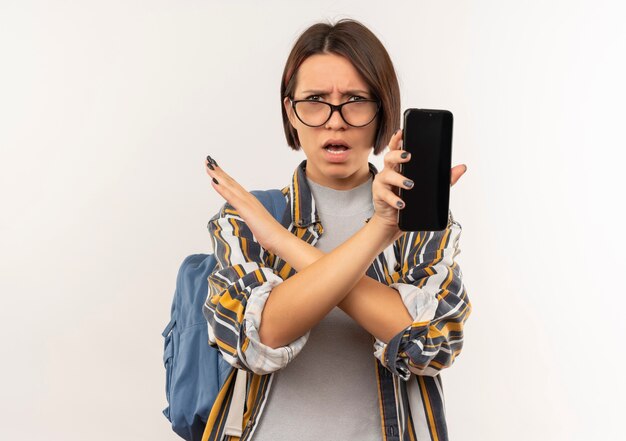 Ragazza giovane studente dispiaciuto con gli occhiali e borsa posteriore che mostra il telefono cellulare non facendo alcun gesto a lato isolato su priorità bassa bianca con lo spazio della copia