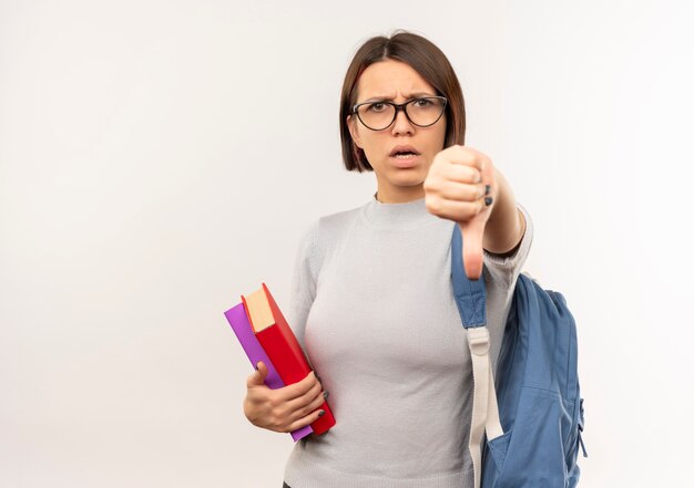 Ragazza giovane studente dispiaciuto con gli occhiali e borsa posteriore che tengono i libri che mostrano il pollice verso il basso isolato su priorità bassa bianca con lo spazio della copia