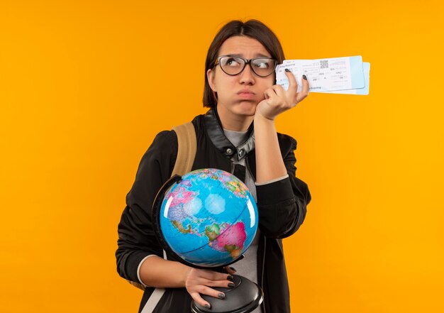 Ragazza giovane studente dispiaciuto con gli occhiali e borsa posteriore che tiene i biglietti aerei e il globo guardando il lato guardando il lato isolato su sfondo arancione con spazio di copia