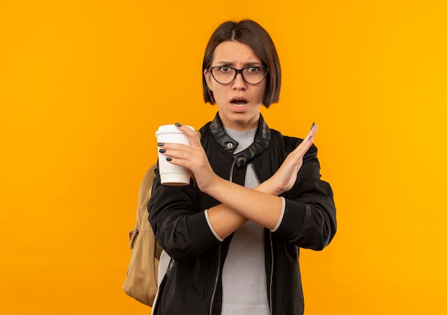 Ragazza giovane studente dispiaciuto con gli occhiali e borsa posteriore che tiene tazza di caffè che gesturing nessun isolato su fondo arancio con lo spazio della copia