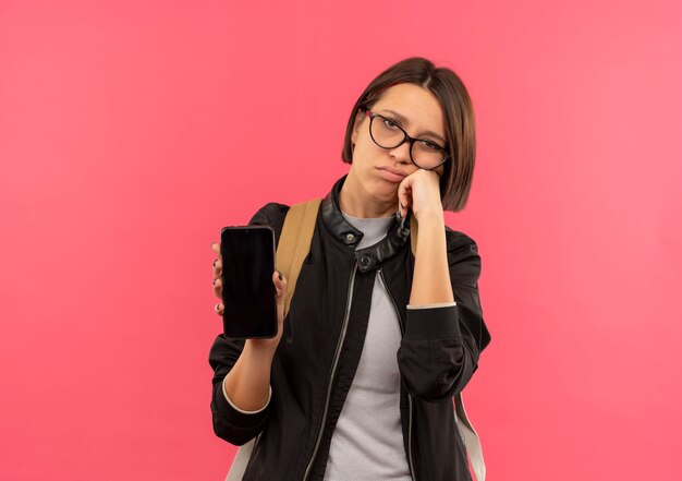 Ragazza giovane studente dispiaciuto con gli occhiali e borsa posteriore tenendo il telefono cellulare mettendo la mano sulla guancia isolata su sfondo rosa con spazio di copia