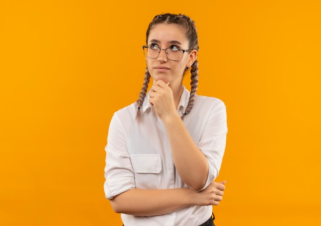 Ragazza giovane studente in bicchieri con le trecce in camicia bianca che osserva da parte con la mano sul mento con espressione pensierosa sul pensiero del viso in piedi sopra la parete arancione