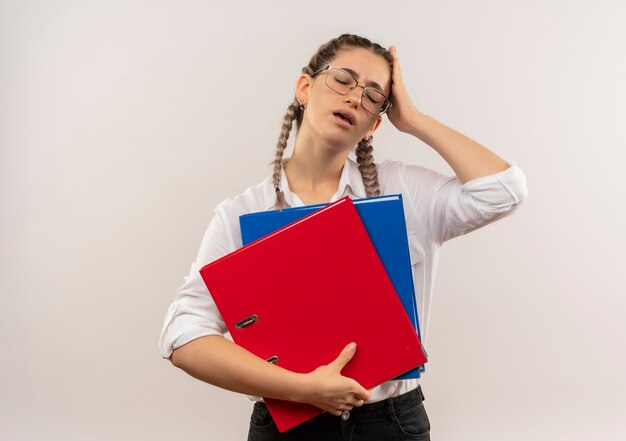 Ragazza giovane studente in bicchieri con le trecce in camicia bianca che tengono le cartelle che sembrano stanchi e oberati di lavoro in piedi sul muro bianco