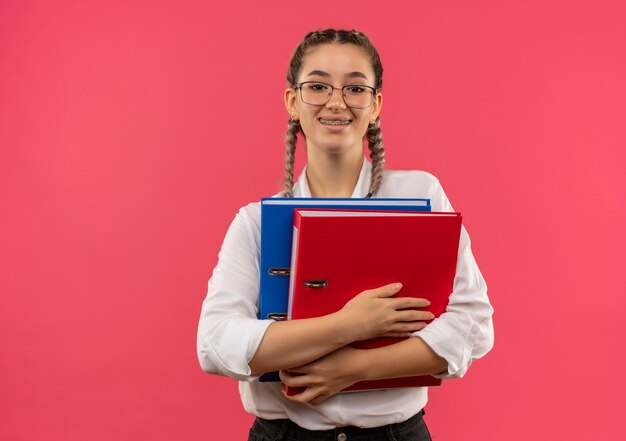 Ragazza giovane studente in bicchieri con le trecce in camicia bianca che tiene due cartelle guardando in avanti sorridendo allegramente in piedi sopra la parete rosa