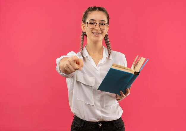 Ragazza giovane studente in bicchieri con le trecce in camicia bianca che tiene il libro puntato con il dito verso la parte anteriore sorridente in piedi sopra la parete rosa