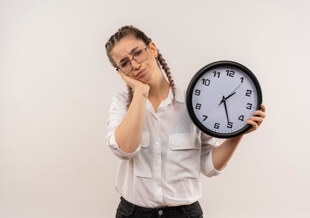 Ragazza giovane studente in bicchieri con le trecce in camicia bianca che tiene l'orologio da parete guardando in avanti stanco e annoiato in piedi sopra il muro bianco