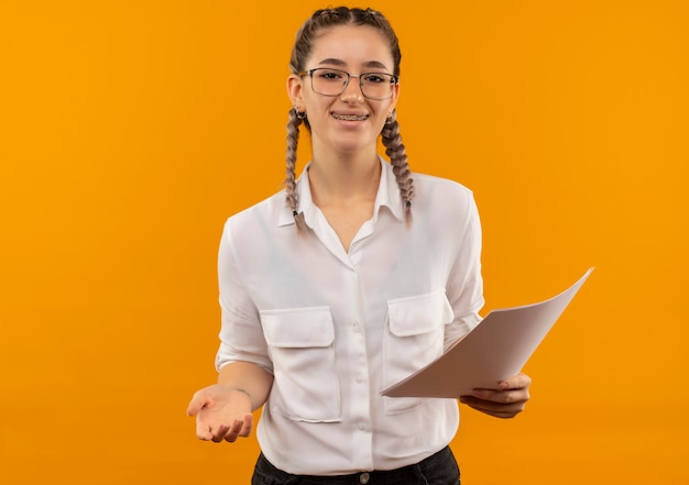 Ragazza giovane studente in bicchieri con le trecce in camicia bianca che tiene pagine vuote guardando in avanti sorridendo allegramente in piedi sopra la parete arancione