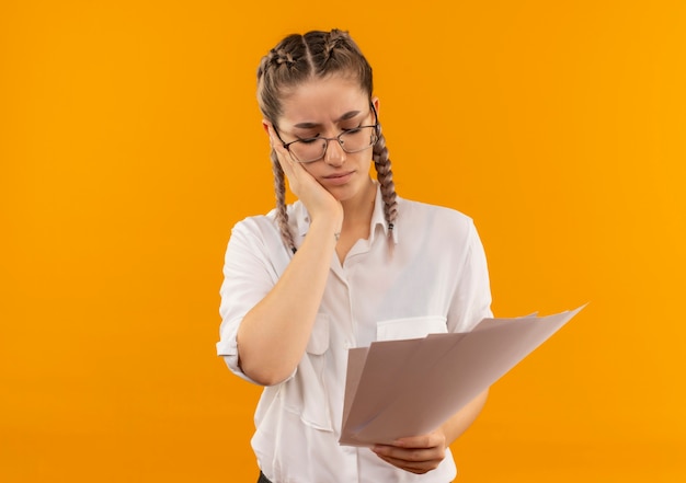 Ragazza giovane studente in bicchieri con le trecce in camicia bianca che tiene pagine vuote guardandoli stanchi e annoiati in piedi sopra la parete arancione