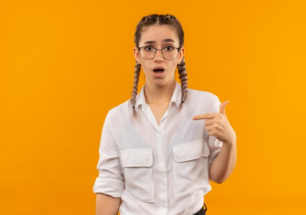 Ragazza giovane studente in bicchieri con le trecce in camicia bianca guardando in avanti confuso che punta a se stessa in piedi sopra la parete arancione