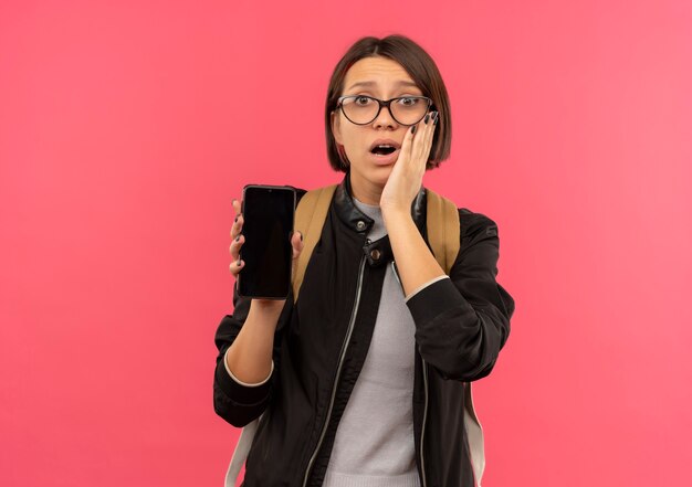 Ragazza giovane studentessa sorpresa con gli occhiali e borsa posteriore che tiene il telefono cellulare mettendo la mano sul viso isolato sulla parete rosa