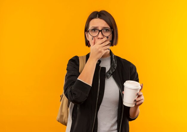 Ragazza giovane studentessa sorpresa con gli occhiali e borsa posteriore che tiene la tazza di caffè di plastica che mette la mano sulla bocca isolata sulla parete arancio