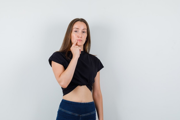 Ragazza giovane tenendo il dito vicino alla bocca in camicetta nera, pantaloni e guardando pensieroso