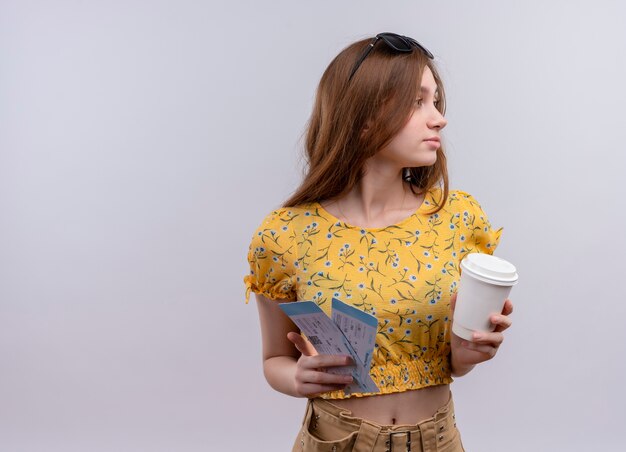 Ragazza giovane viaggiatore indossando occhiali da sole sulla testa che tiene i biglietti aerei e la tazza di caffè di plastica e guardando i biglietti in piedi in vista di profilo sul muro bianco isolato