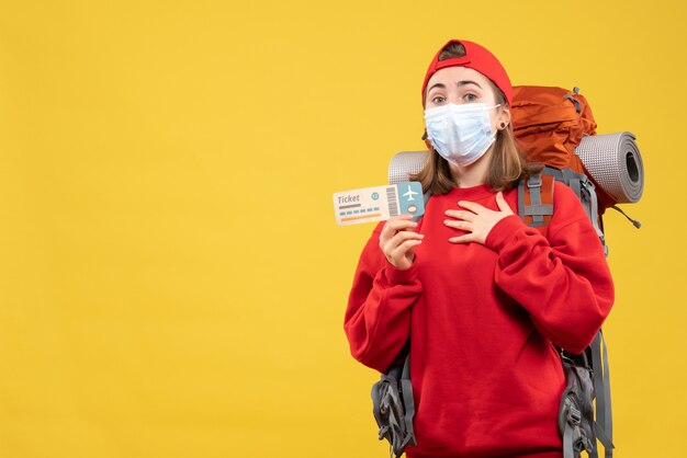 Ragazza giovane viaggiatore vista frontale con zaino e maschera tenendo il biglietto mettendo la mano sul petto