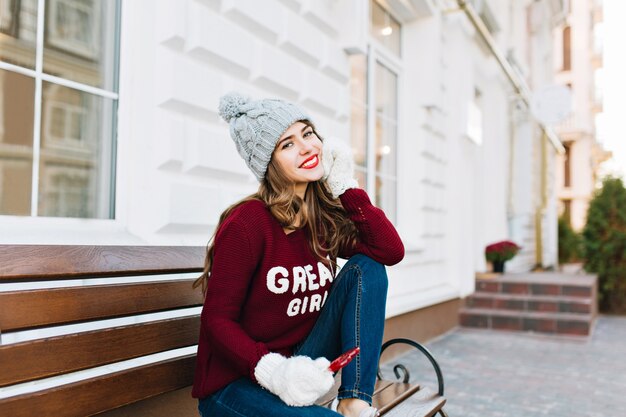 Ragazza graziosa con capelli lunghi in cappello invernale, jeans e guanti bianchi che si siede sulla panchina sulla strada. Ha conseguito il cuore rosso caramello, sorridendo.