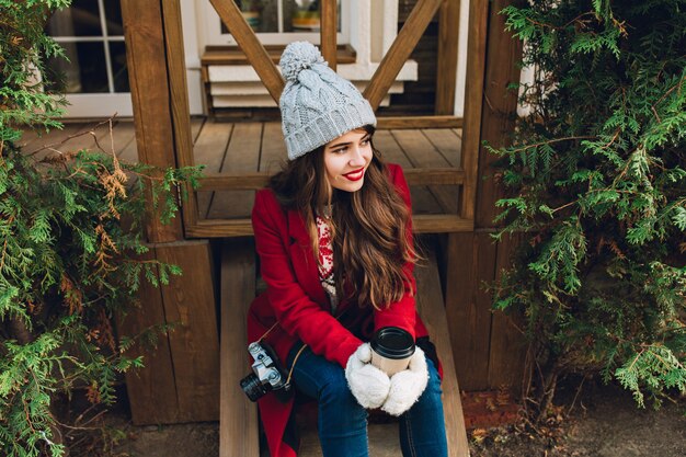 Ragazza graziosa con capelli lunghi in cappotto rosso che si siede sulle scale di legno tra rami verdi all'aperto. Ha un cappello lavorato a maglia grigio, tiene il caffè in guanti bianchi e sorride a lato.