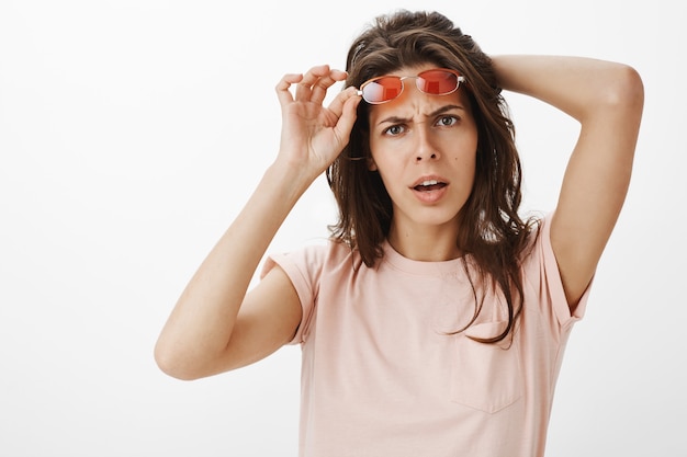 Ragazza graziosa confusa con gli occhiali da sole che posano contro il muro bianco