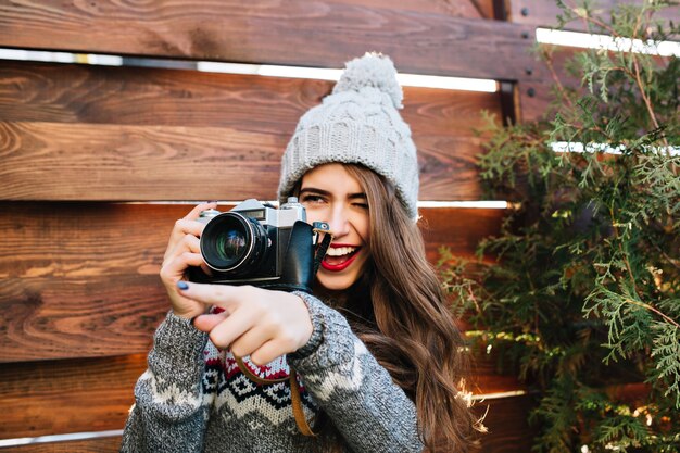 Ragazza graziosa del ritratto con capelli lunghi in cappello lavorato a maglia divertendosi a fare una foto sulla macchina fotografica su legno.